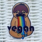Light brown sloth vomitting a rainbow with "vegan" across the front sticker image displayed on white basket background