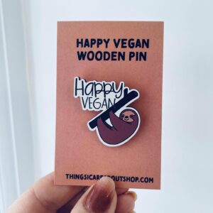 Hand holding up a cardboard backing with the Happy Vegan sloth wooden pin which is a smiling sloth holding onto a stick and the words Happy Vegan above the stick.