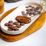 Oat milk chocolate buttons on a white ceramic dish on top of a wooden platter, with some of the chocolate melted with a metal spoon under the melted chocolate.